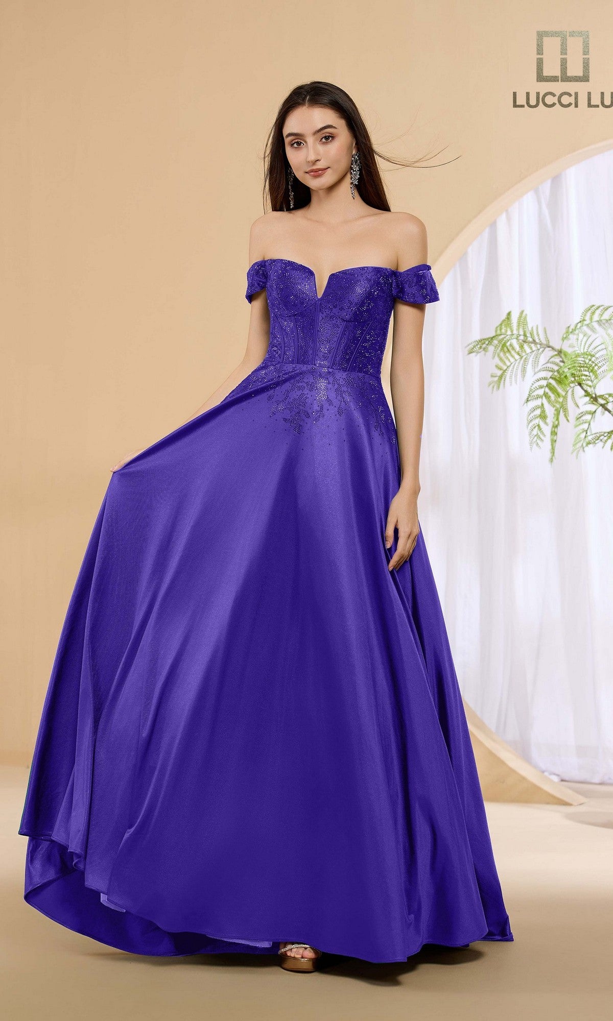 A woman wears the Lucci Lu 1483 long purple prom dress, featuring a vibrant off-the-shoulder stretch satin design with a corset bodice and flared skirt, while standing indoors against a beige background.
